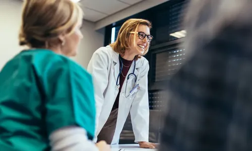 Smiling female healthcare educator in a white coat with a stethoscope leaning on a table, engaging with nursing students during a classroom discussion Banner Smiling female nurse educator in a white coat with a stethoscope leaning on a table, engaging with nursing students during a classroom discussion