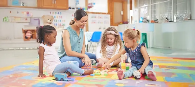 Preschool teacher interacting with a group of young children playing with building blocks on a colorful mat in a classroom setting.