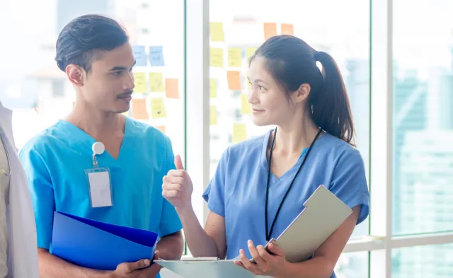 Two nurses are talking and looking at each other in their blue scrubs. They're holding binders.