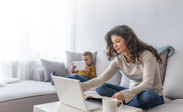 Mother seated on couch taking an online class with son reading on a tablet next to her