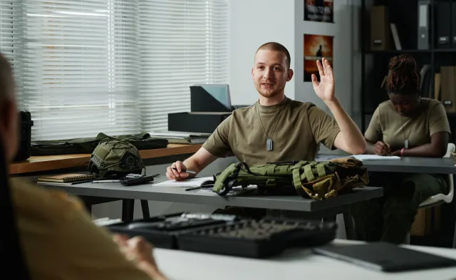 Young male student raising hand and looking at teacher at lesson while asking him question after lecture about military training