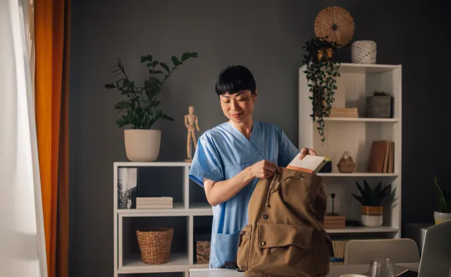 Asian dedicated nurse in blue scrubs is seen preparing medical tools and organizing documents into a brown backpack in a well-lit, modern room with shelves and assorted decor.