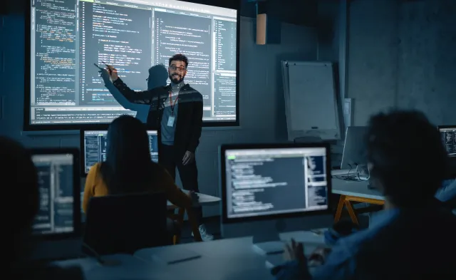 Teacher Giving Computer Science Lecture to Diverse Multiethnic Group of Female and Male Students in Dark College Room. Projecting Slideshow with Programming Code. Explaining Information Technology.