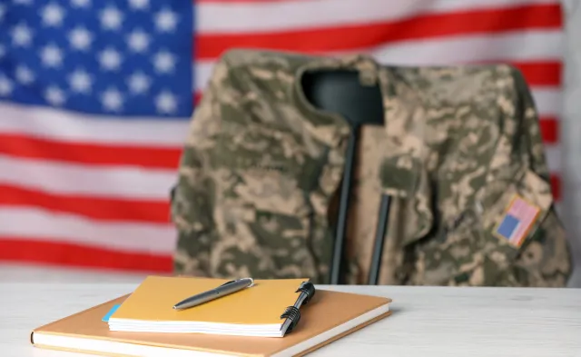 Notebooks and pen on white wooden table, chair with soldier uniform against flag of United States indoors. Military education