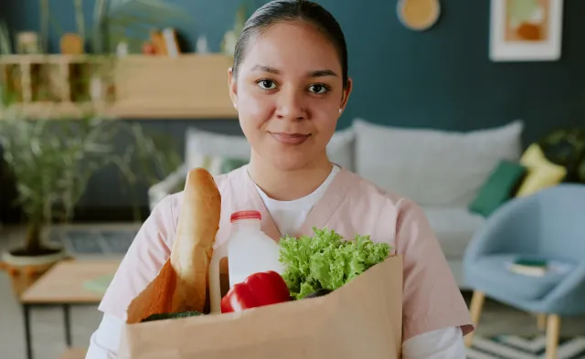 Young nurse holding paper bag full of healthy food while looking at camera and smiling widely
