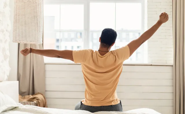 Arabic Man Sitting On Bed Back To Camera And Stretching Arms Awaking After Good Sleep In Modern Bedroom At Home, Looking Out Of Window. Comfort And Coziness. Good Morning Concept
