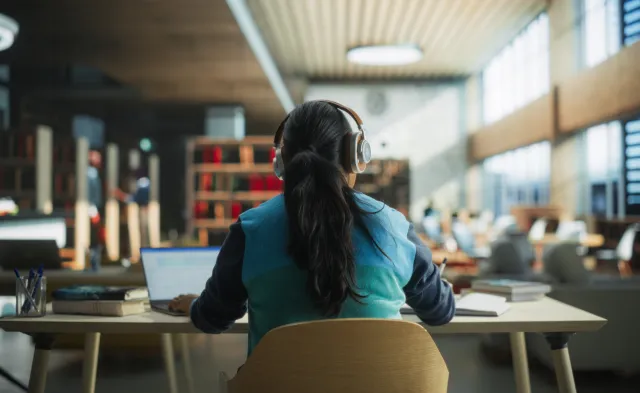 Female Student Wearing Headphones while Working on University Homework in a Public Library. Woman Sitting Behind a Desk, Using Laptop Computer and Writing Down Notes in Notebook. Footage from the Back