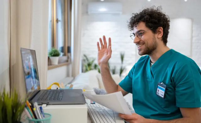 A young Caucasian male nurse in scrubs, having a video call while working from his home