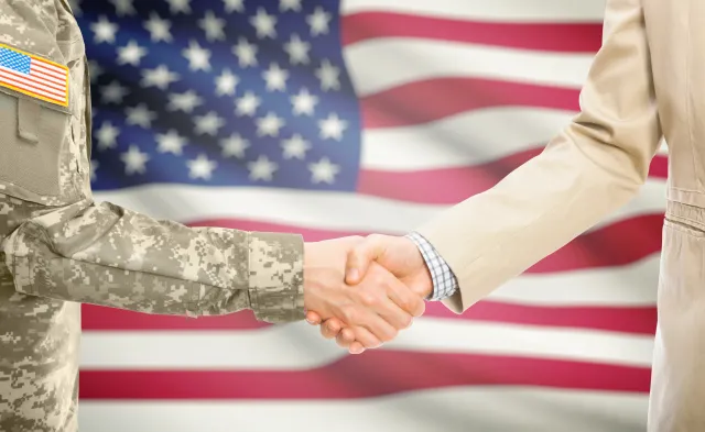 American soldier in uniform and civil man in suit shaking hands with national flag on background - United States