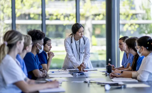 Nursing instructor in a white lab coat leads a group discussion with nursing students seated around a table in a bright classroom, reviewing medical charts and notes during a team learning session