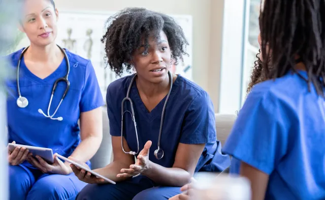 Nurses in blue scrubs sit together in a team meeting, discussing patient care while holding tablets.