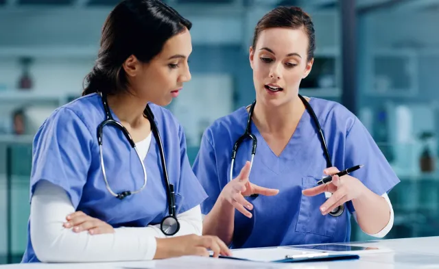 Two nurses in blue scrubs discuss treatment notes at a desk with a tablet and clipboard.