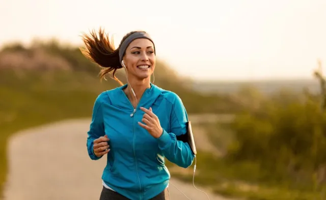 Woman smiling while jogging outdoors on a winding path, wearing earbuds and a blue jacket with a phone armband.