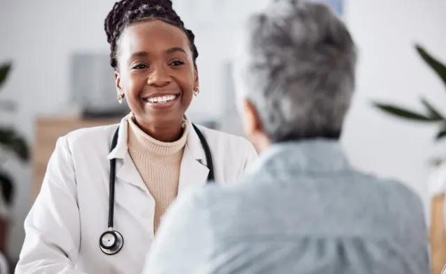 A smiling nurse practitioner wearing a white coat and stethoscope speaks with an older patient during an appointment.