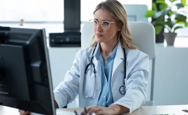 A nurse practitioner wearing a white lab coat and stethoscope sits at a desk using a computer in a bright medical office.