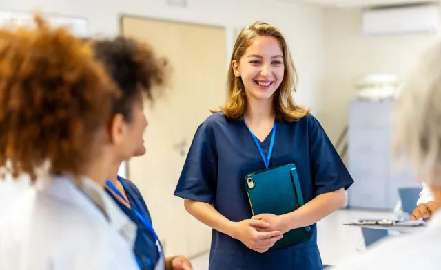 Smiling nursing student in scrubs holding a tablet while speaking with healthcare professionals in a clinical setting.