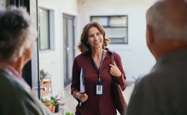 Home healthcare professional wearing an ID badge stands at a front door smiling and speaking with two older adults while holding a clipboard, suggesting an in-home wellness or care visit.
