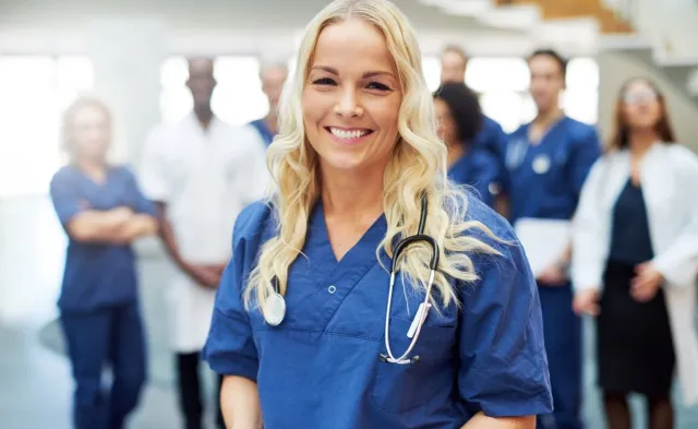 Smiling RN who is pursuing a BSN in blue scrubs with a stethoscope stands in the foreground while a multidisciplinary medical team gathers in the background of a healthcare facility.