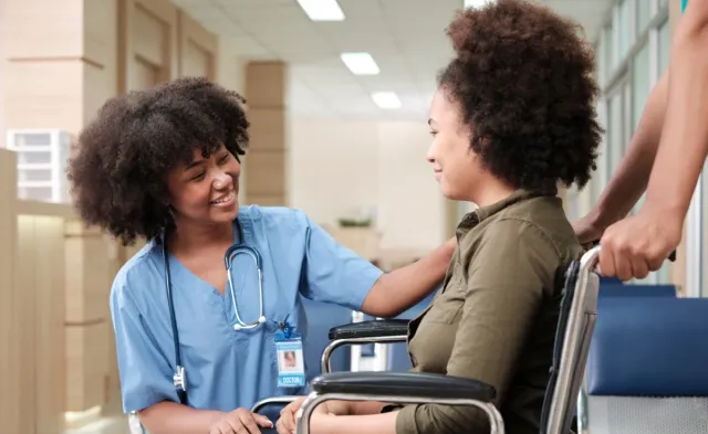 Registered nurse in blue scrubs speaks reassuringly with a patient seated in a wheelchair in a hospital hallway.