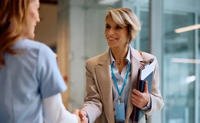 Professional shakes hands with a healthcare worker while holding a clipboard, suggesting a meeting or career opportunity in a healthcare setting.