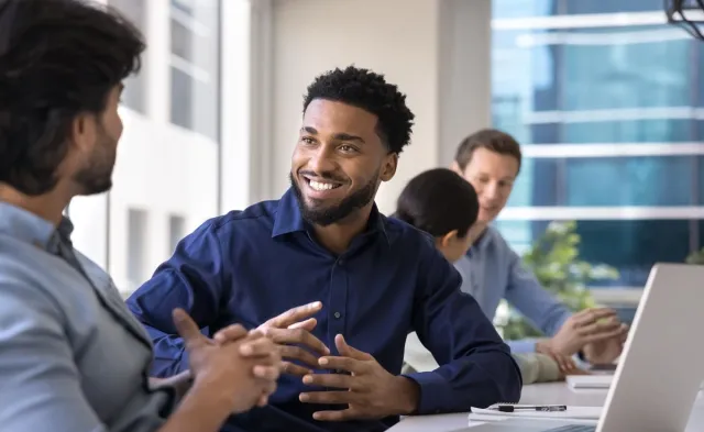 Four coworkers sit around a table in a bright office. A smiling man in a navy shirt talks with a colleague in the foreground while two others work near an open laptop in the background.