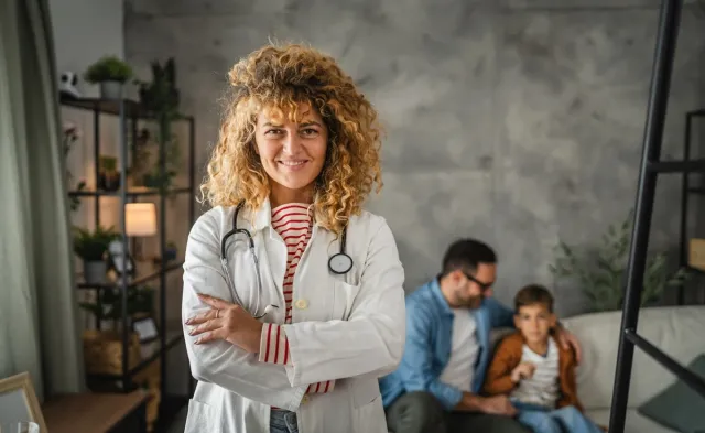 Smiling FNP in a white coat and stethoscope standing with arms crossed while an adult and child sit together in the background.
