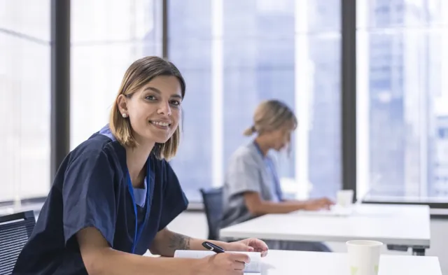 A nursing student studying at a table writes notes in a notebook while another student works quietly in the background near large windows.