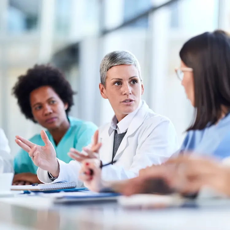 A nurse executive with both an MSN and MBA in a white coat facilitates a collaborative team meeting with nurses and doctors in a hospital conference room.