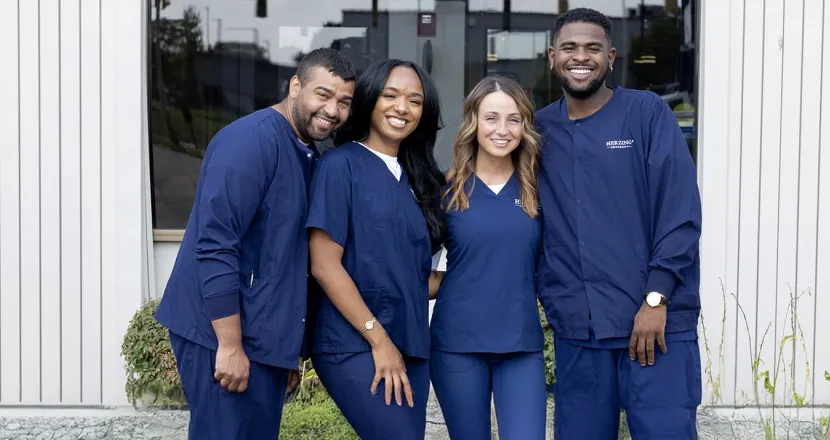 Wide-angle group photo of Herzing University medical assisting students smiling in navy scrubs outside a campus building, emphasizing teamwork and inclusivity.