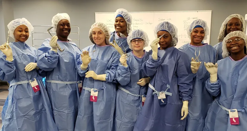 A group of surgical technology students in scrubs, holding medical instruments and smiling in a classroom setting, showcasing teamwork and learning.
