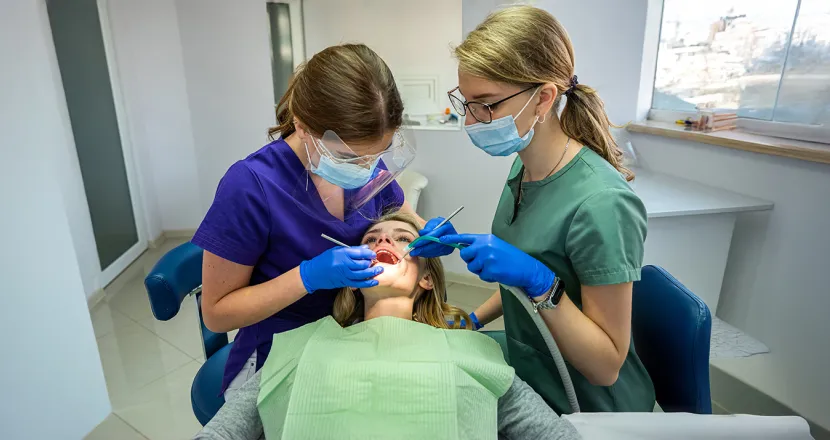 Two dental professionals in protective gear and masks performing a dental procedure on a patient in a modern dental office, showcasing teamwork and hygiene practices.