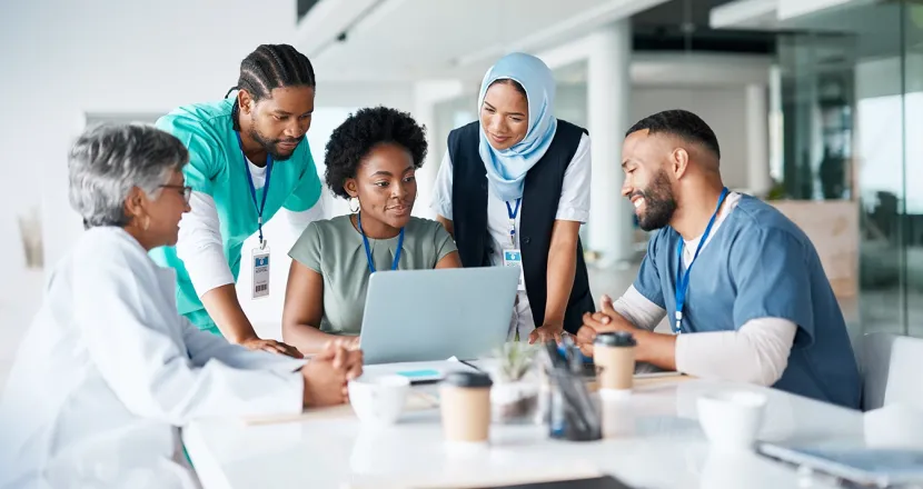 Diverse healthcare professionals collaborating around a laptop during a meeting, demonstrating teamwork and leadership in healthcare administration.