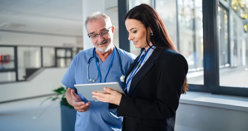 Healthcare professional in scrubs discussing information with a businesswoman in a blazer, both looking at a tablet in a modern office setting.