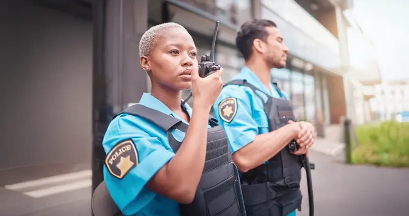 Two uniformed officers, a woman using a walkie-talkie and a man standing beside her, maintain a professional stance outside a modern building, representing the career paths available to criminal justice graduates.