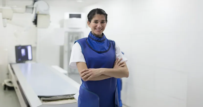 Radiologic technologist standing confidently in a clinical setting wearing a lead apron near medical imaging equipment.