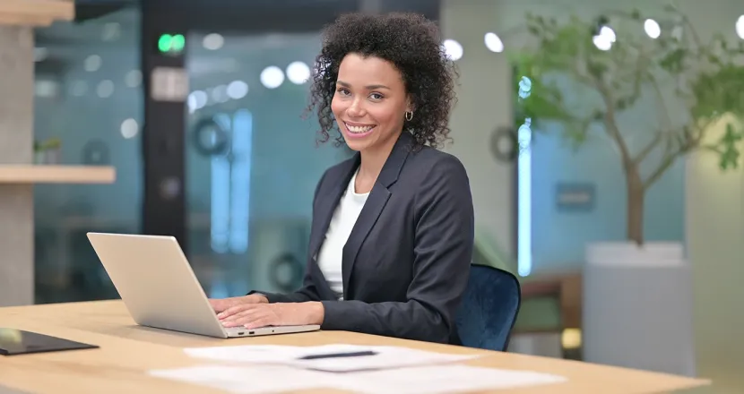 A professional woman sits at a desk with a laptop, smiling confidently, representing career success with a business management degree.