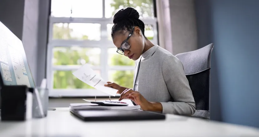 A Herzing University accounting student in professional attire reviews financial documents at a modern office desk, preparing reports and analyzing data.