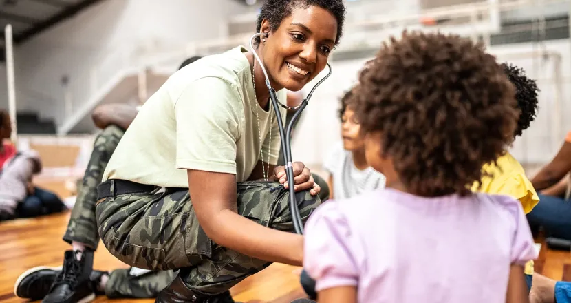 Smiling military service member using a stethoscope to check a young child’s heartbeat, demonstrating community healthcare and support.