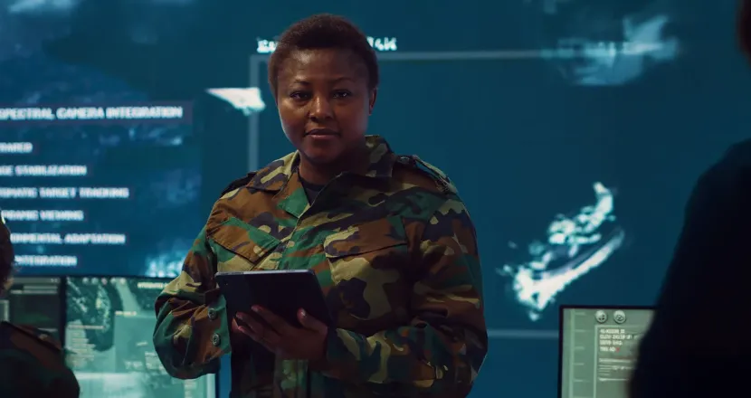 Military service member in camouflage uniform holding a tablet in a high-tech operations center with digital surveillance screens in the background.