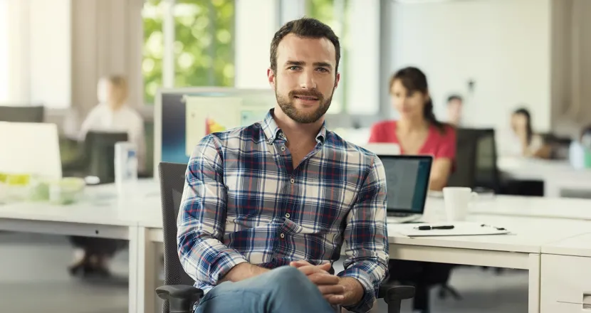 A Herzing University interdisciplinary studies student in a casual plaid shirt sits confidently in a modern, collaborative office environment, ready for a versatile career path.