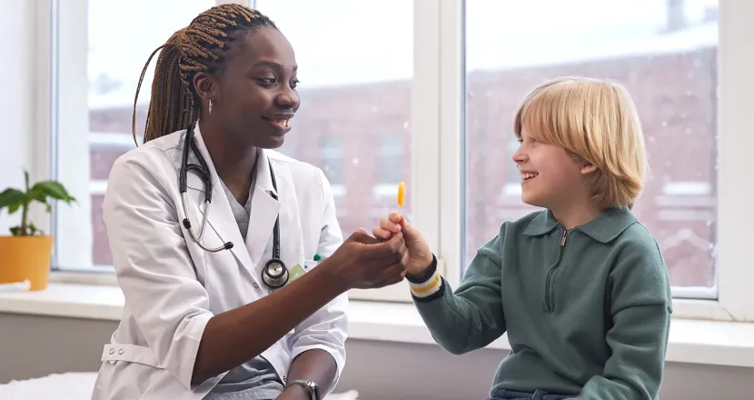 Woman healthcare professional smiling and handing a lollipop to a young boy in a clinic setting, with snow visible outside the window