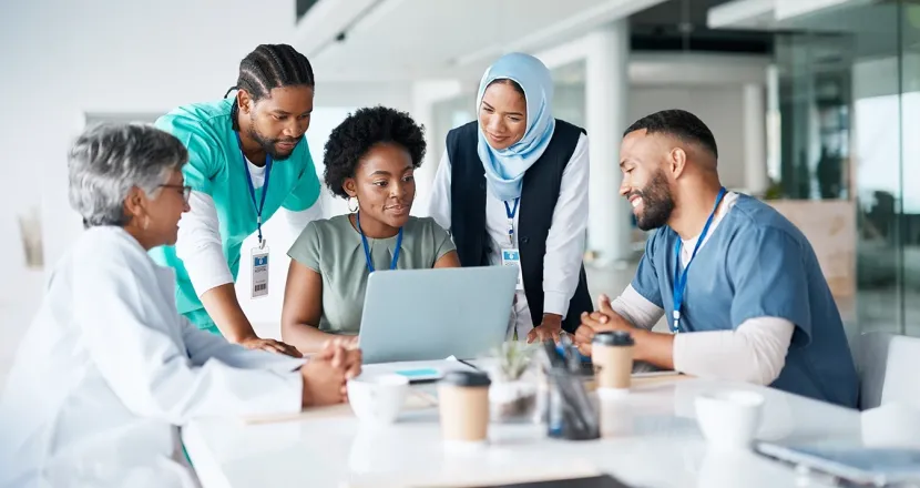 Diverse healthcare professionals collaborating around a laptop during a meeting, demonstrating teamwork and leadership in healthcare administration.
