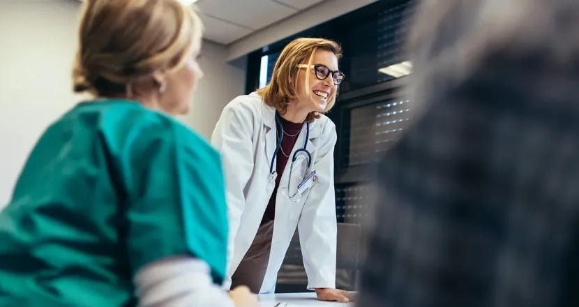 Smiling female healthcare educator in a white coat with a stethoscope leaning on a table, engaging with nursing students during a classroom discussion