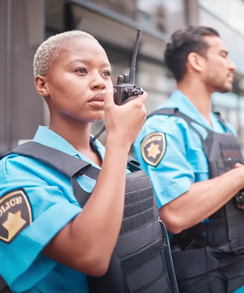 Two uniformed officers, a woman using a walkie-talkie and a man standing beside her, maintain a professional stance outside a modern building, representing the career paths available to criminal justice graduates.