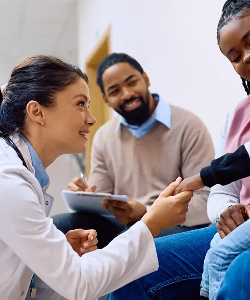 Smiling family nurse practitioner kneeling and greeting a young boy sitting on his mother's lap in a medical waiting area, with the boy's father smiling in the background