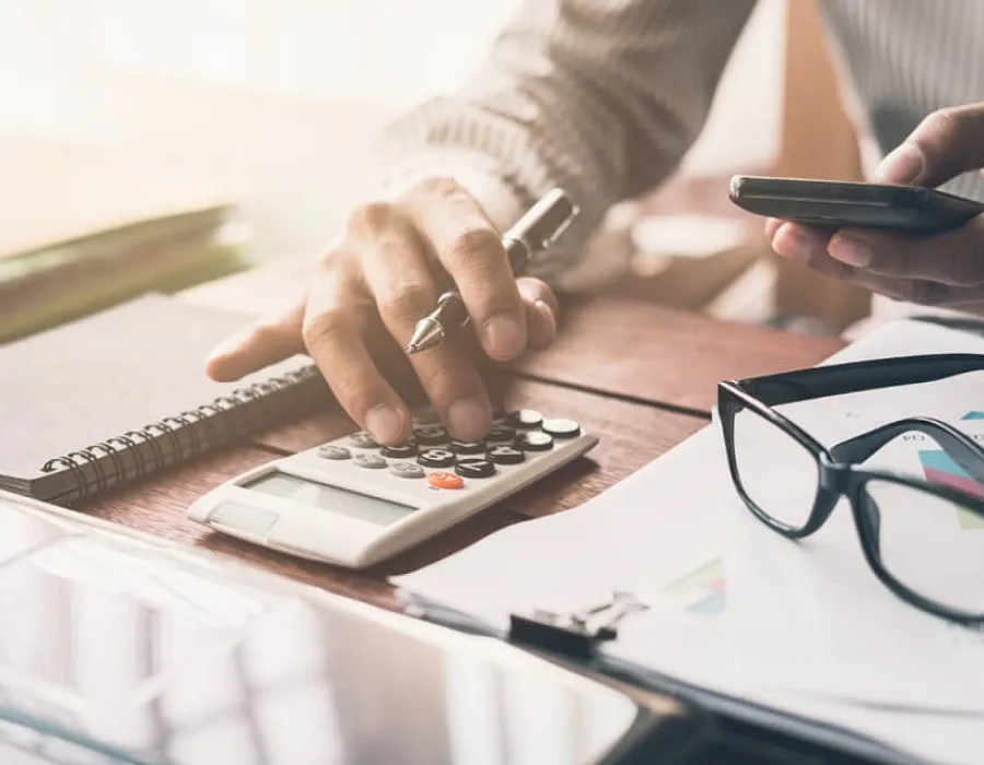 Accountant punching in calculations on calculator with glasses resting on desktop