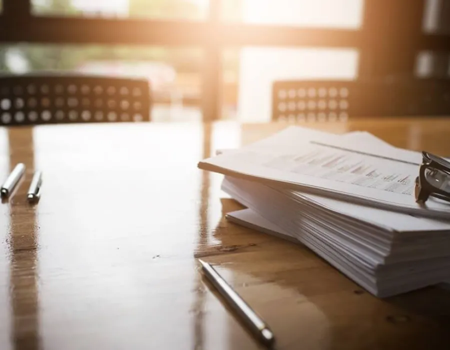 Stack of legal papers on a sunlit desk with glasses rested on top and pens on table nearby