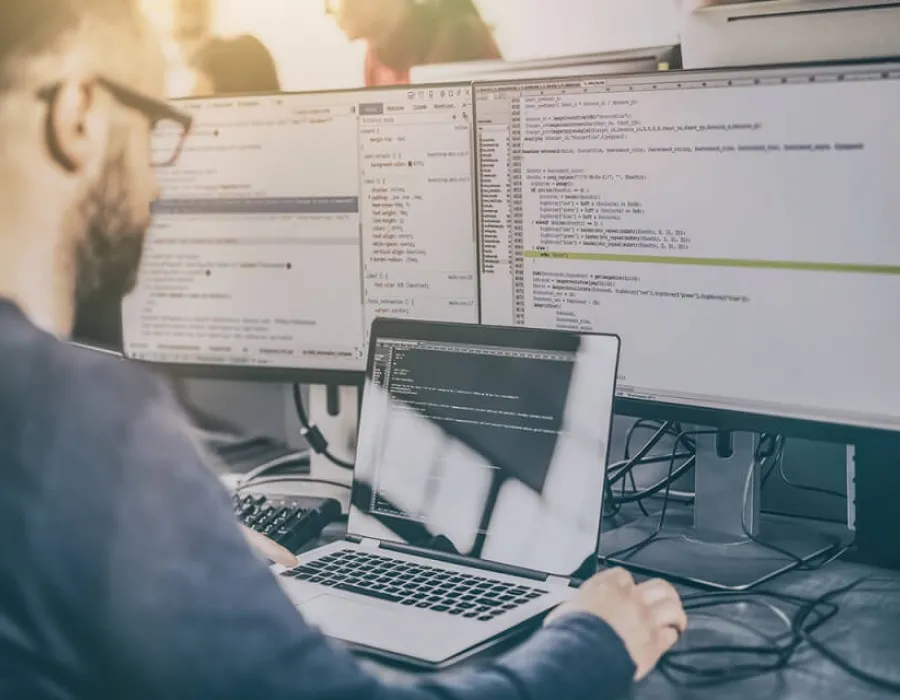 Computer programmer seated in front of two monitors and laptop executing code