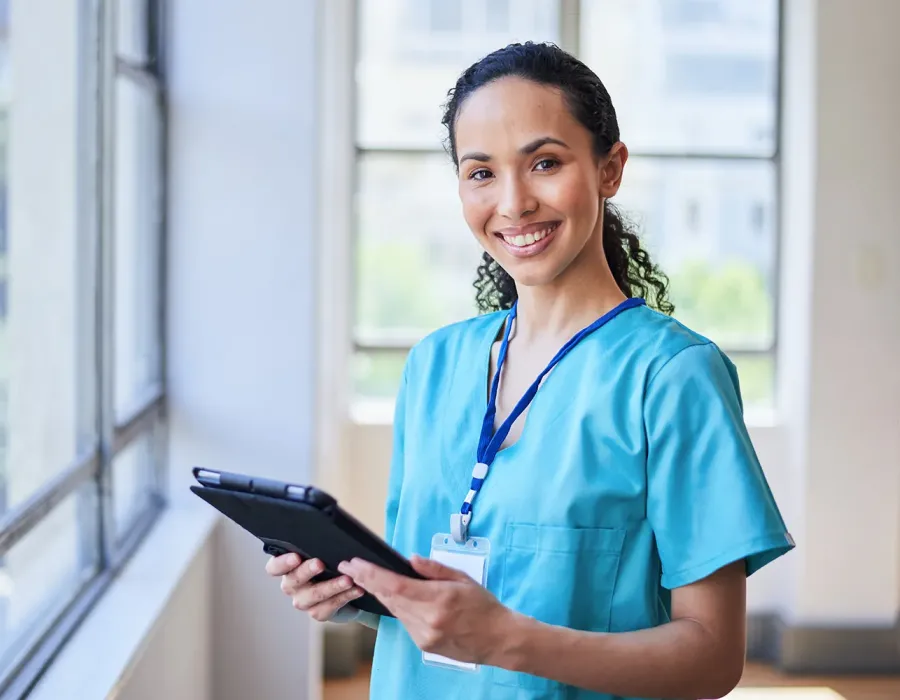 Practical nurse with prior medical assistant experience wearing blue scrubs holding clipboard in medical clinic