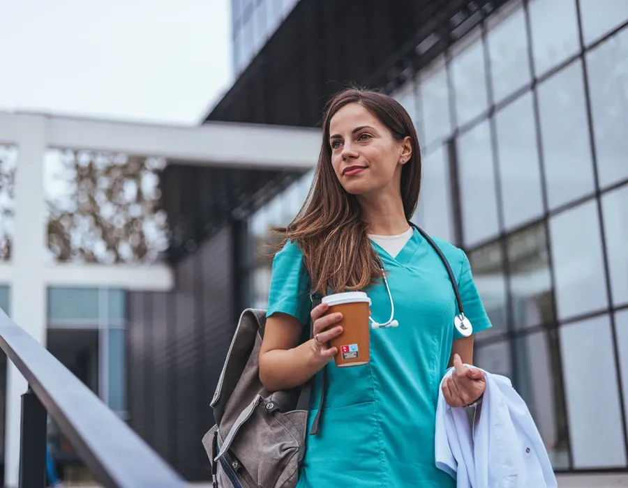 Woman travel nurse wearing green scrubs and backpack bringing coffee to work